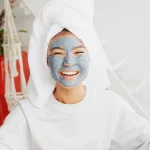 Femme souriante avec une serviette sur la tête et un masque visage hydratant argileux