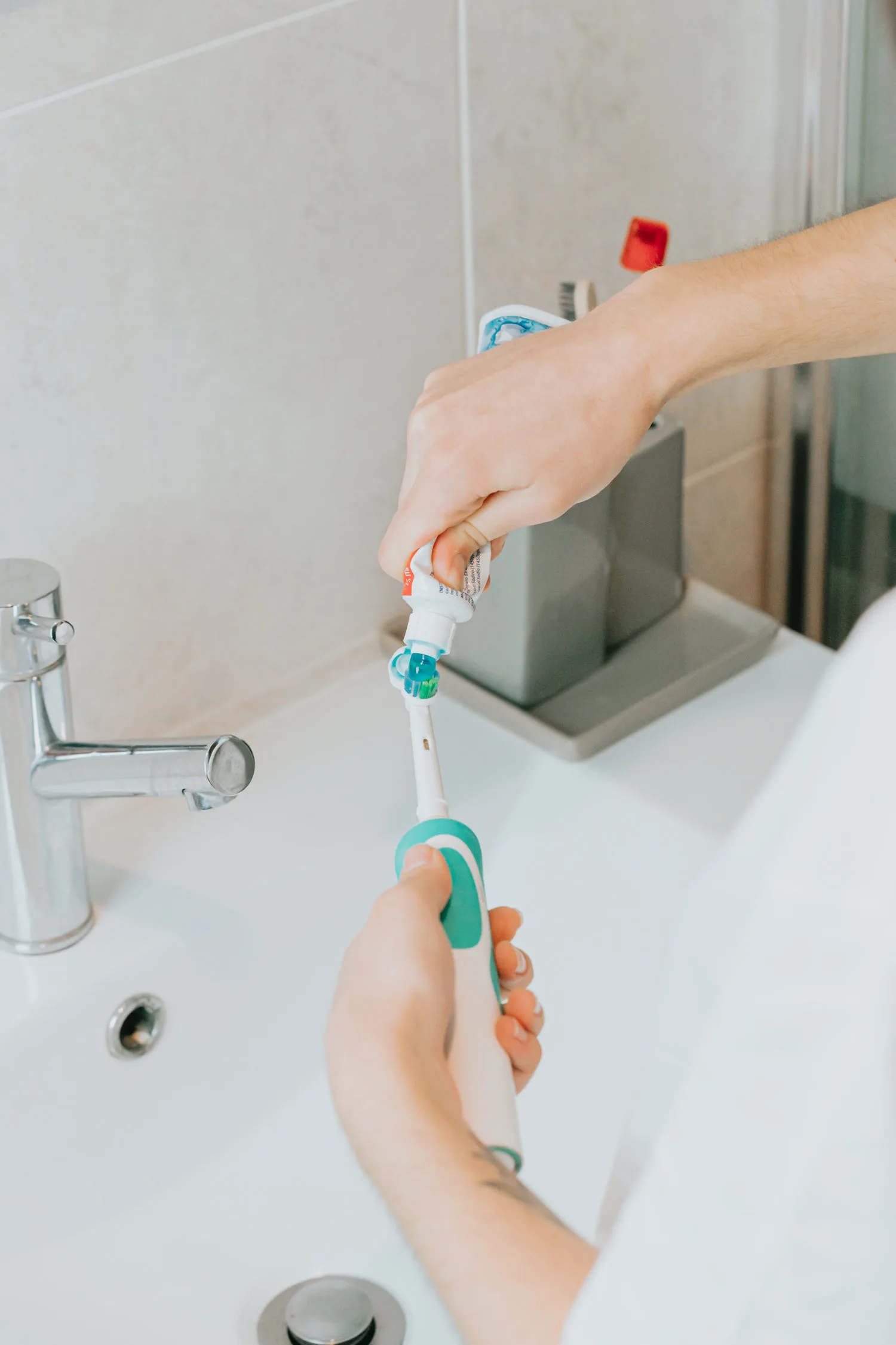 Mains versant du dentifrice sur une brosse à dents électrique au-dessus d'un lavabo blanc