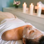 Femme allongée en spa avec masque visage en tissu blanc entourée de bougies blanches